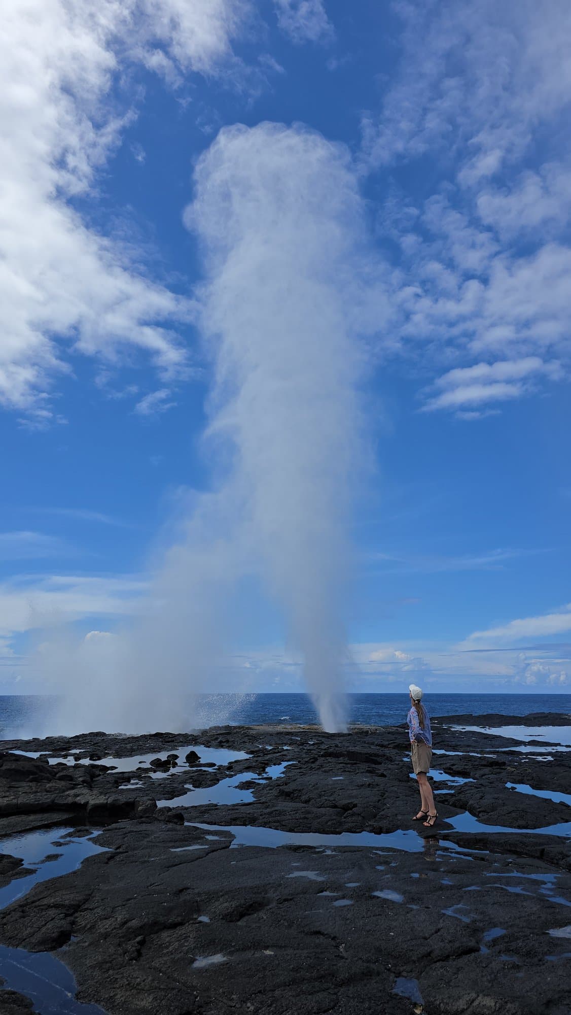 An Alofaaga blowhole firing seawater high above the lava shelf, a person tiny beside it