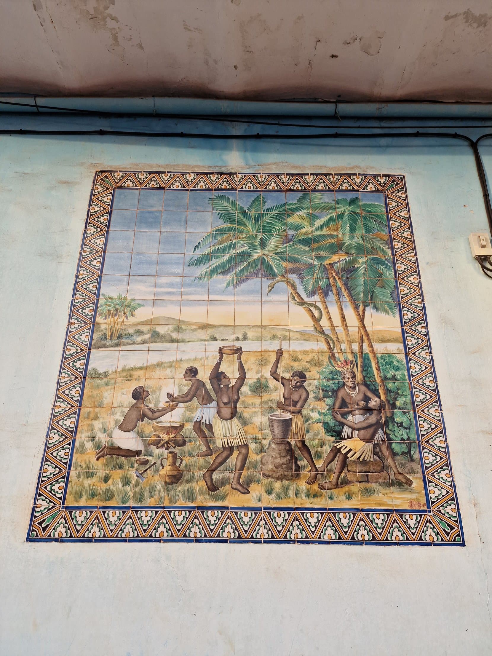 A blue and white Portuguese tile panel showing a harvest scene under palms, on a wall in Bissau