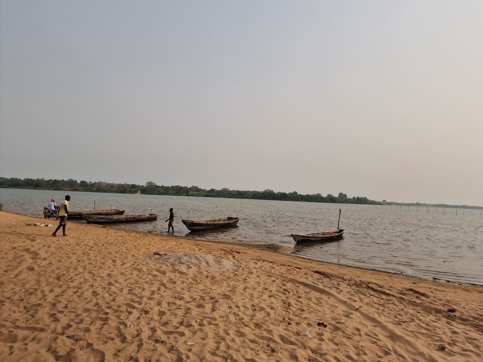 Wooden boats and a few figures on the sandy shore of Lake Togo in flat afternoon light