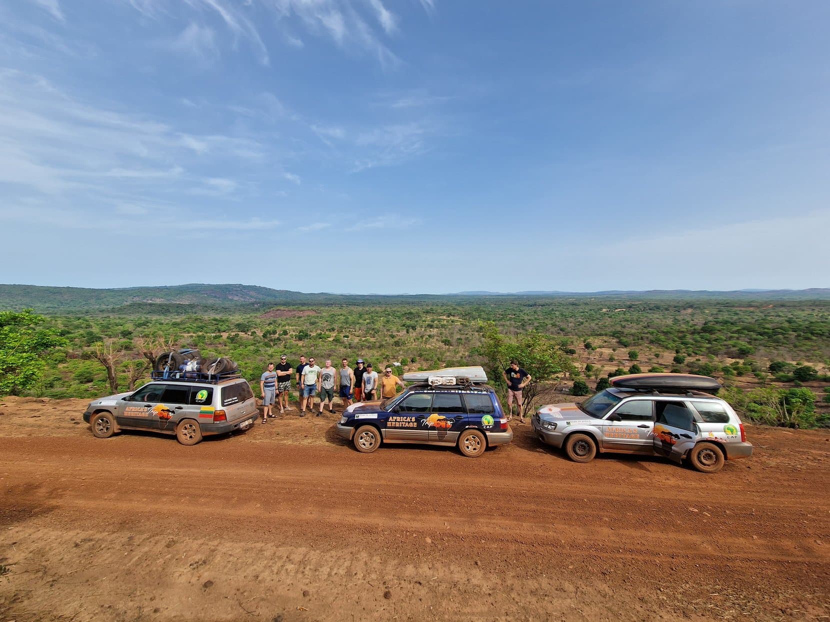 Three rally cars parked on a red dirt road with a long escarpment behind, Guinea