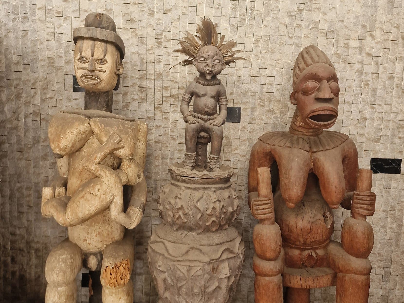 Three large carved wooden figures against a stone wall inside a museum, Lomé, Togo