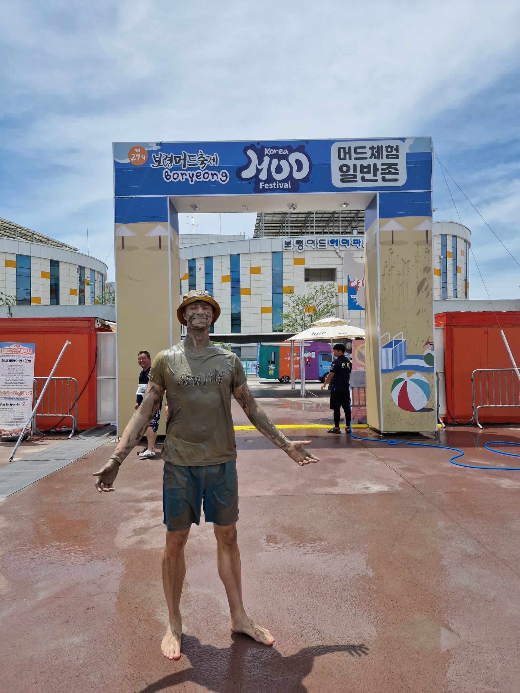 A man covered head to toe in grey mud, arms out, under the entrance arch of the Boryeong Mud Festival
