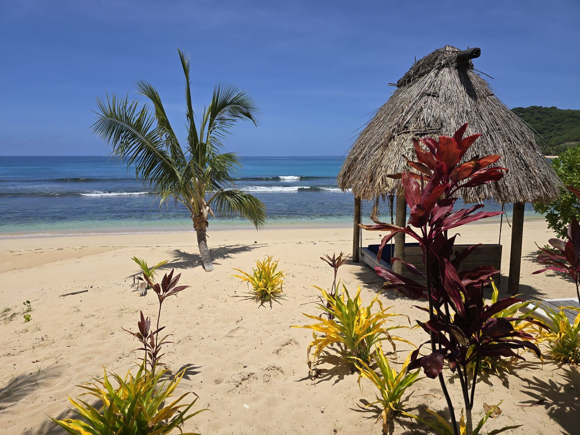 A thatched pavilion and red ti plants on a white-sand island beach in Fiji