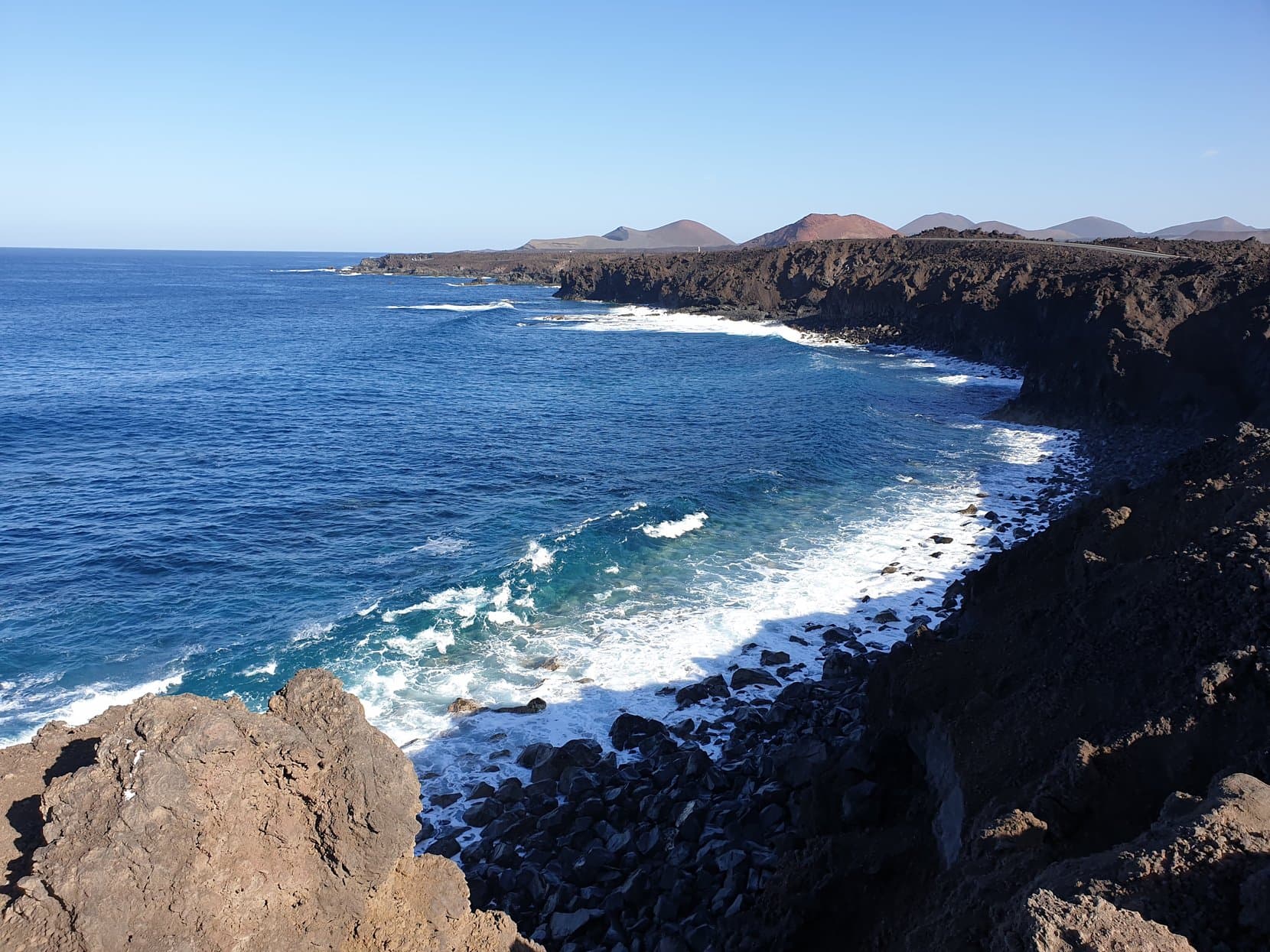 Waves breaking white against the black lava coast of Los Hervideros, volcano cones on the skyline, Lanzarote