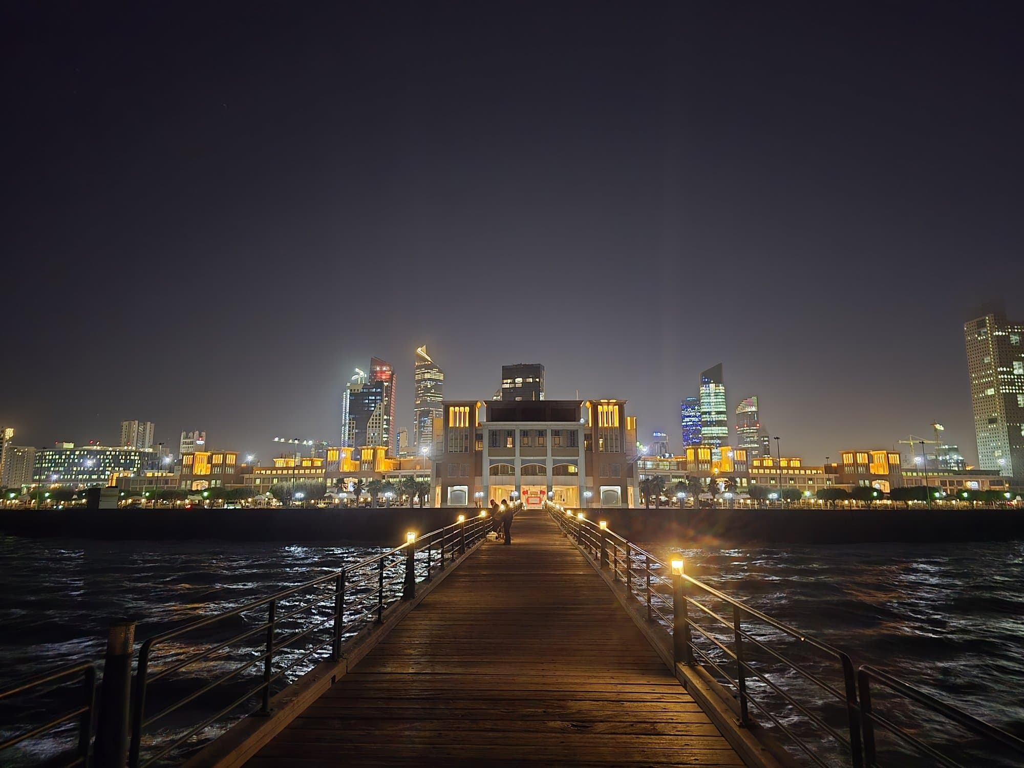 The wooden pier at Souq Sharq marina at night, the lit skyline behind