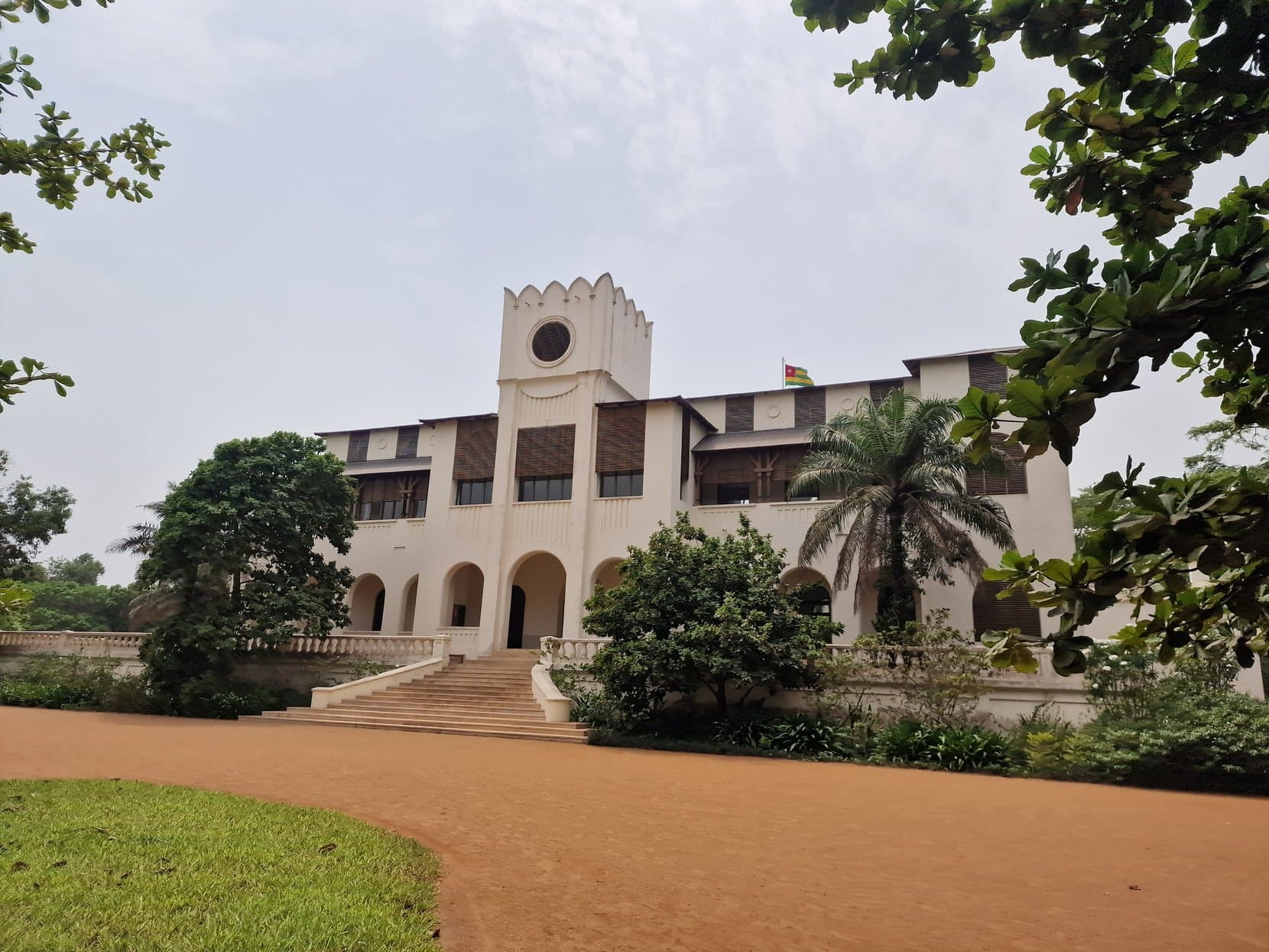 The white facade of the Palais de Lomé behind trees and a sandy drive, Togo