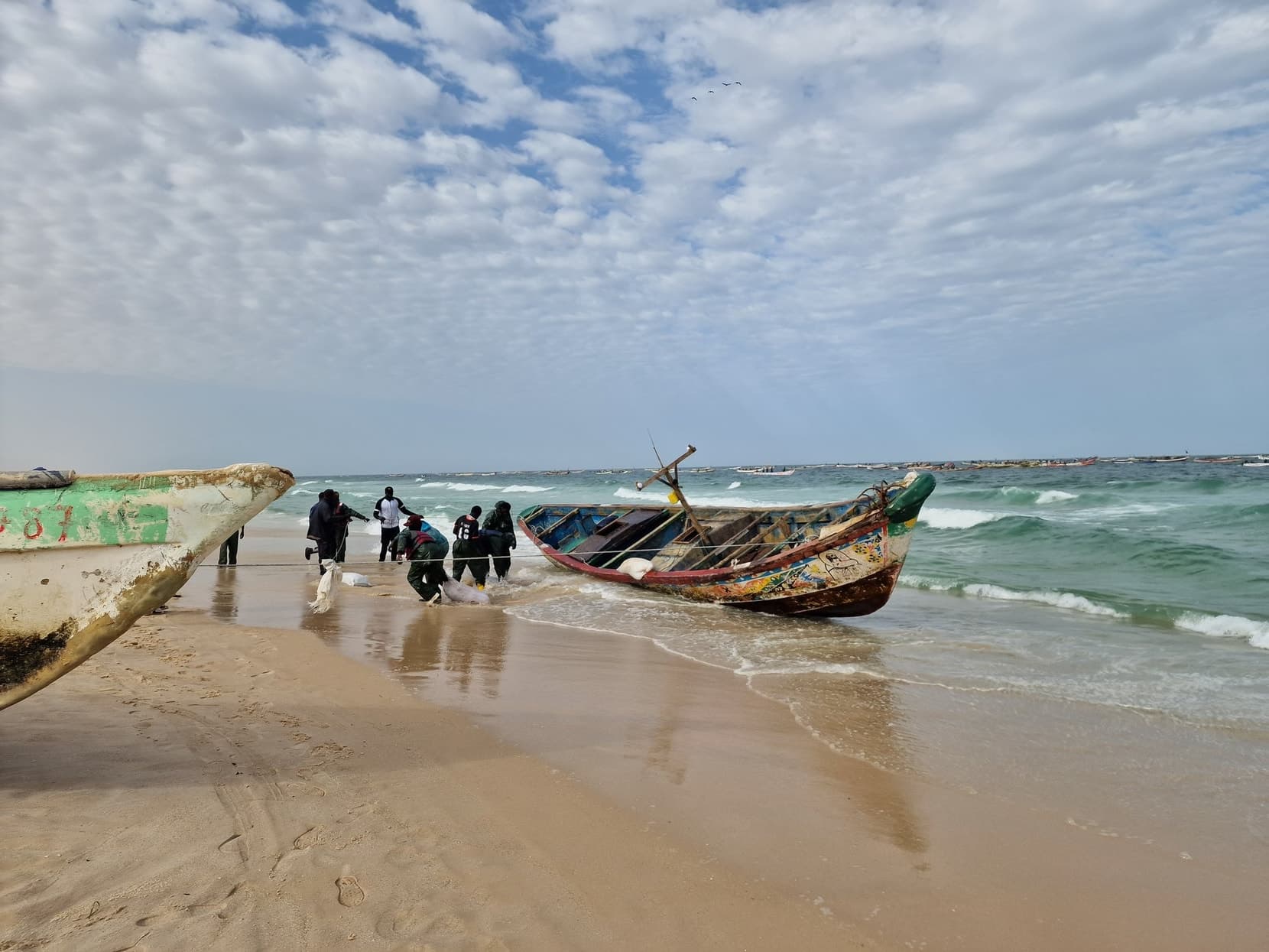 A painted wooden pirogue landing through the surf, fishermen hauling it up the sand