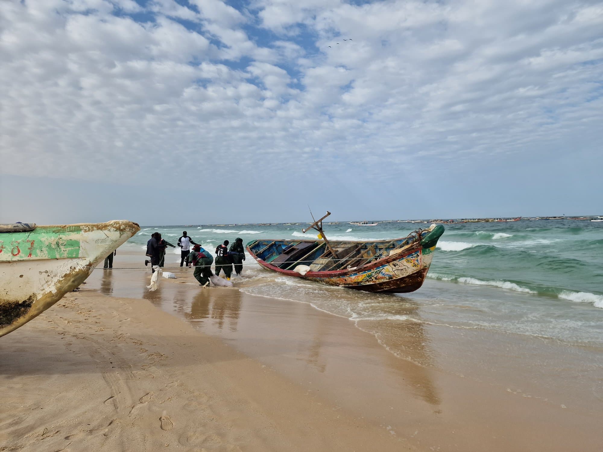 A painted wooden pirogue landing through the surf, fishermen hauling it up the sand