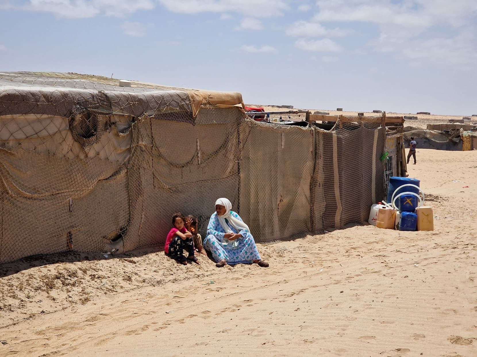 A Sahrawi camp by the beach, figures resting in the shade of a tent wall