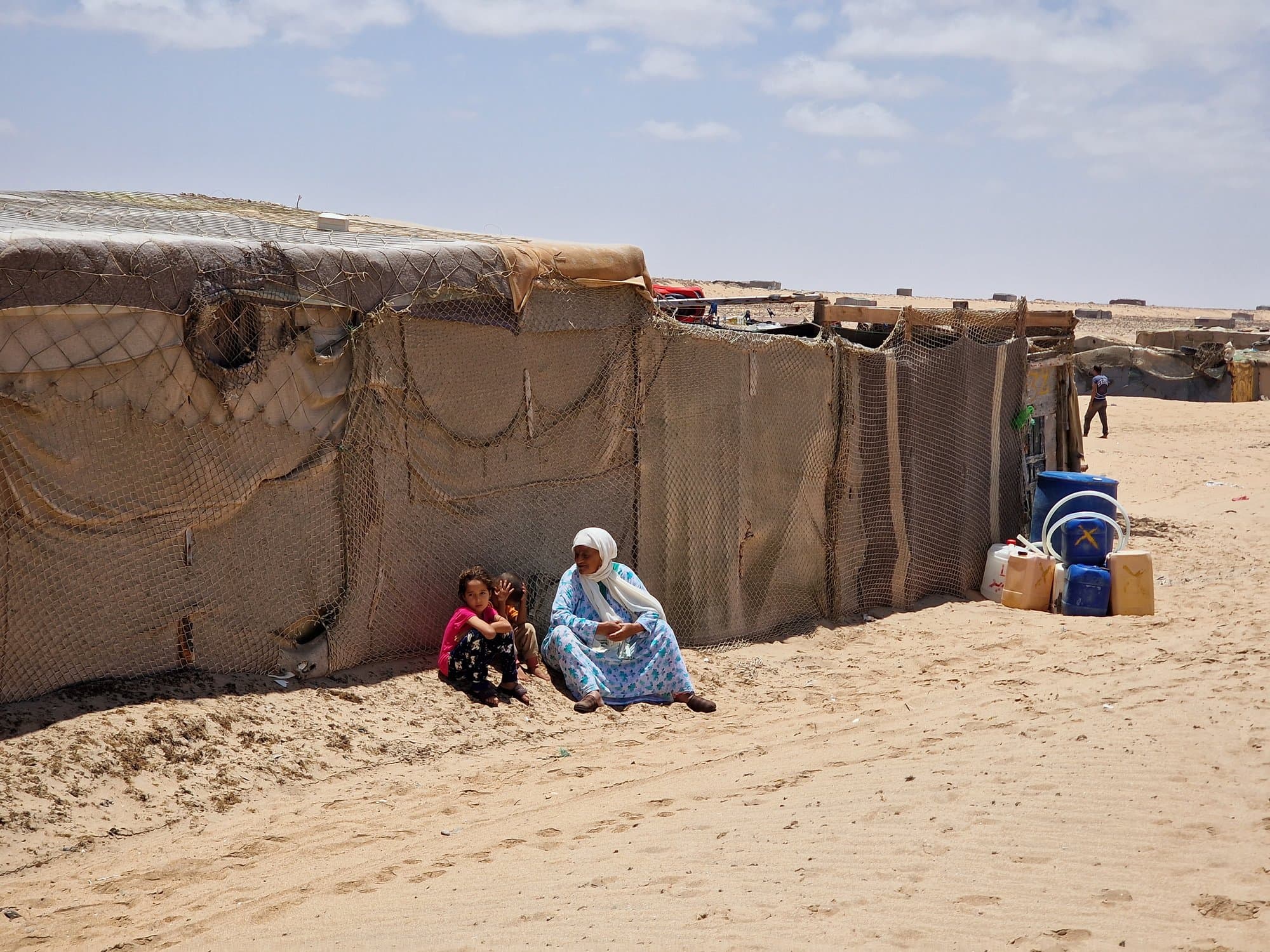 A Sahrawi camp by the beach, figures resting in the shade of a tent wall