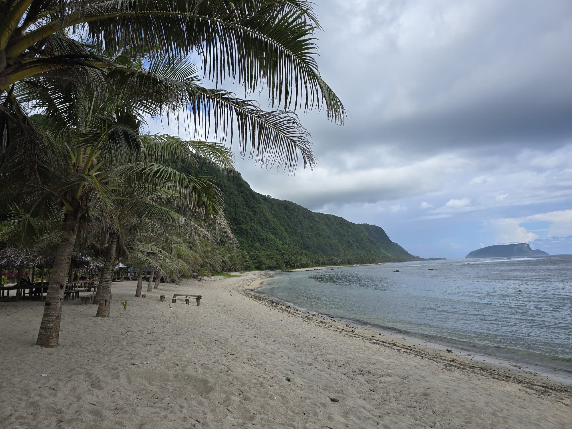 An empty white-sand beach on Upolu's south-east coast, green cliffs behind and an island offshore
