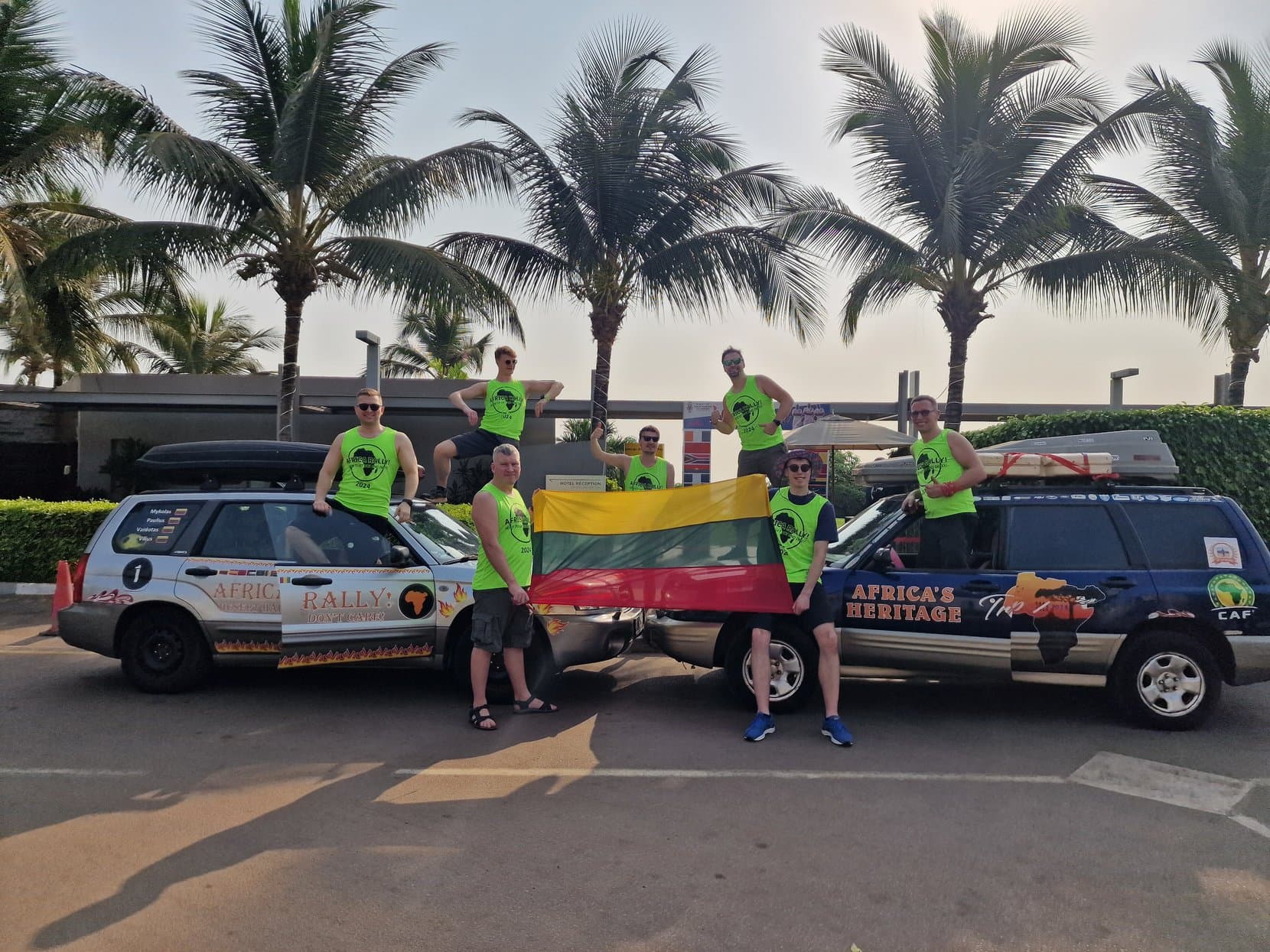 The 2024 rally crew with a Lithuanian flag beside two Subarus under palms, Conakry
