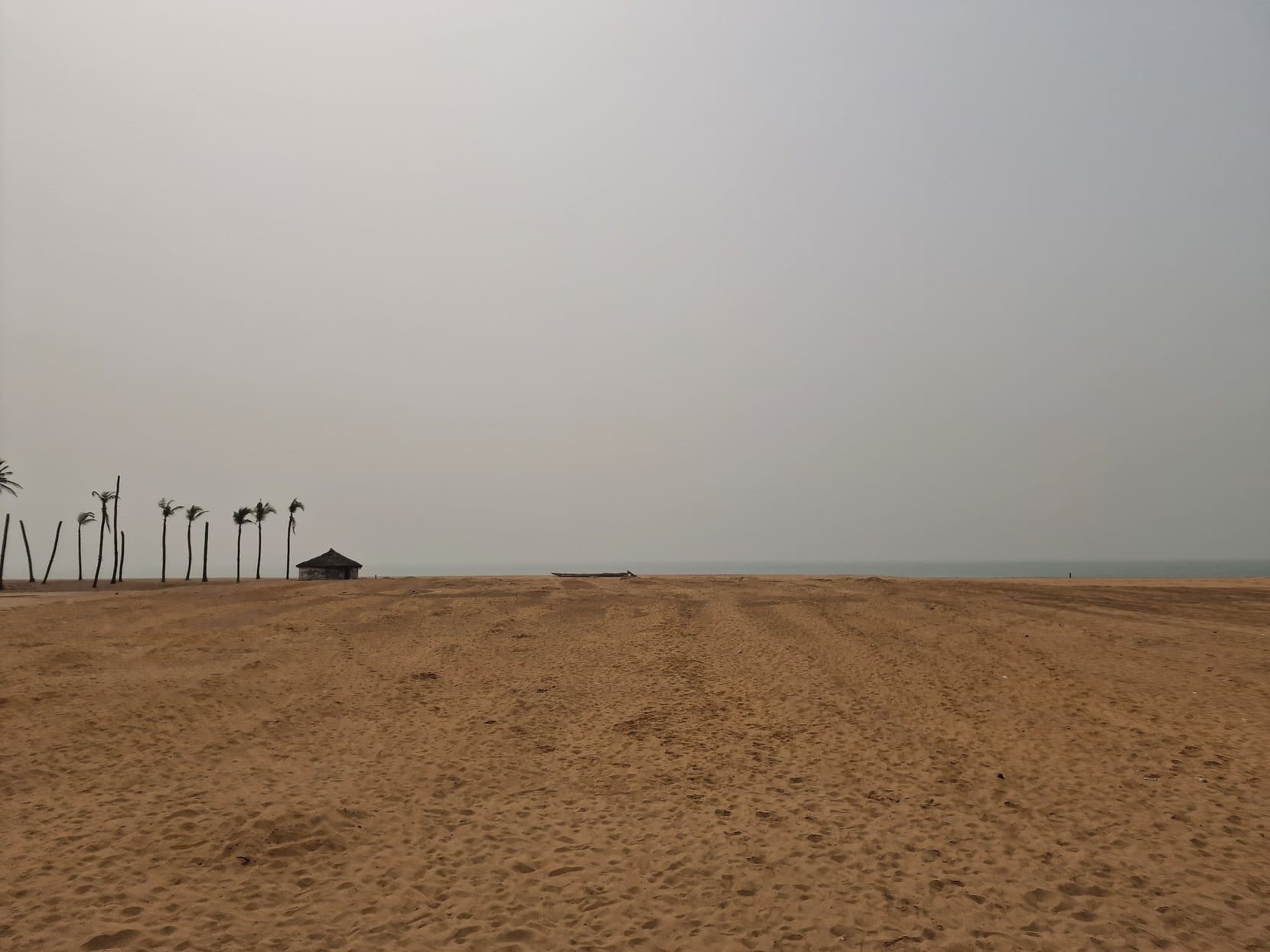 An empty stretch of sand with a single thatched shelter and a few palms fading into haze, the beach at Ouidah, Benin
