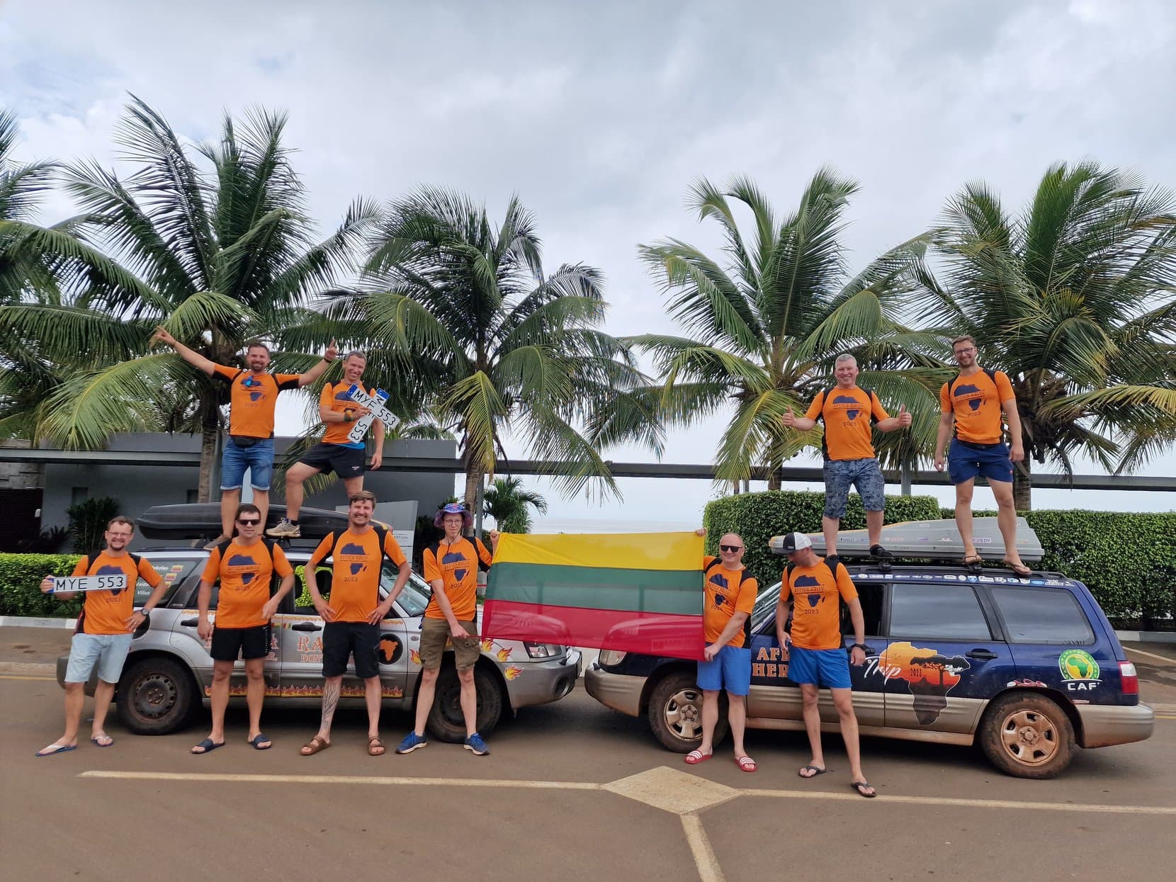 The rally crew and two Subarus with a Lithuanian flag under palms in Conakry, Guinea