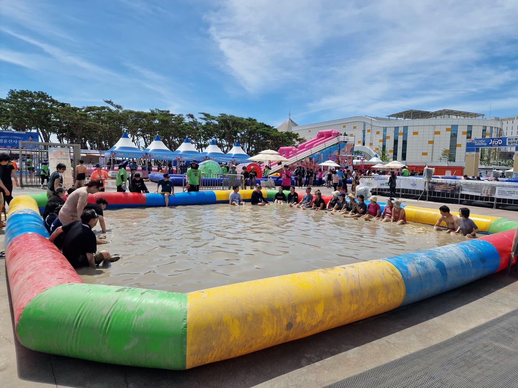 A crowded mud pool ringed by inflatables under a blue sky at the Boryeong Mud Festival, Daecheon Beach, South Korea