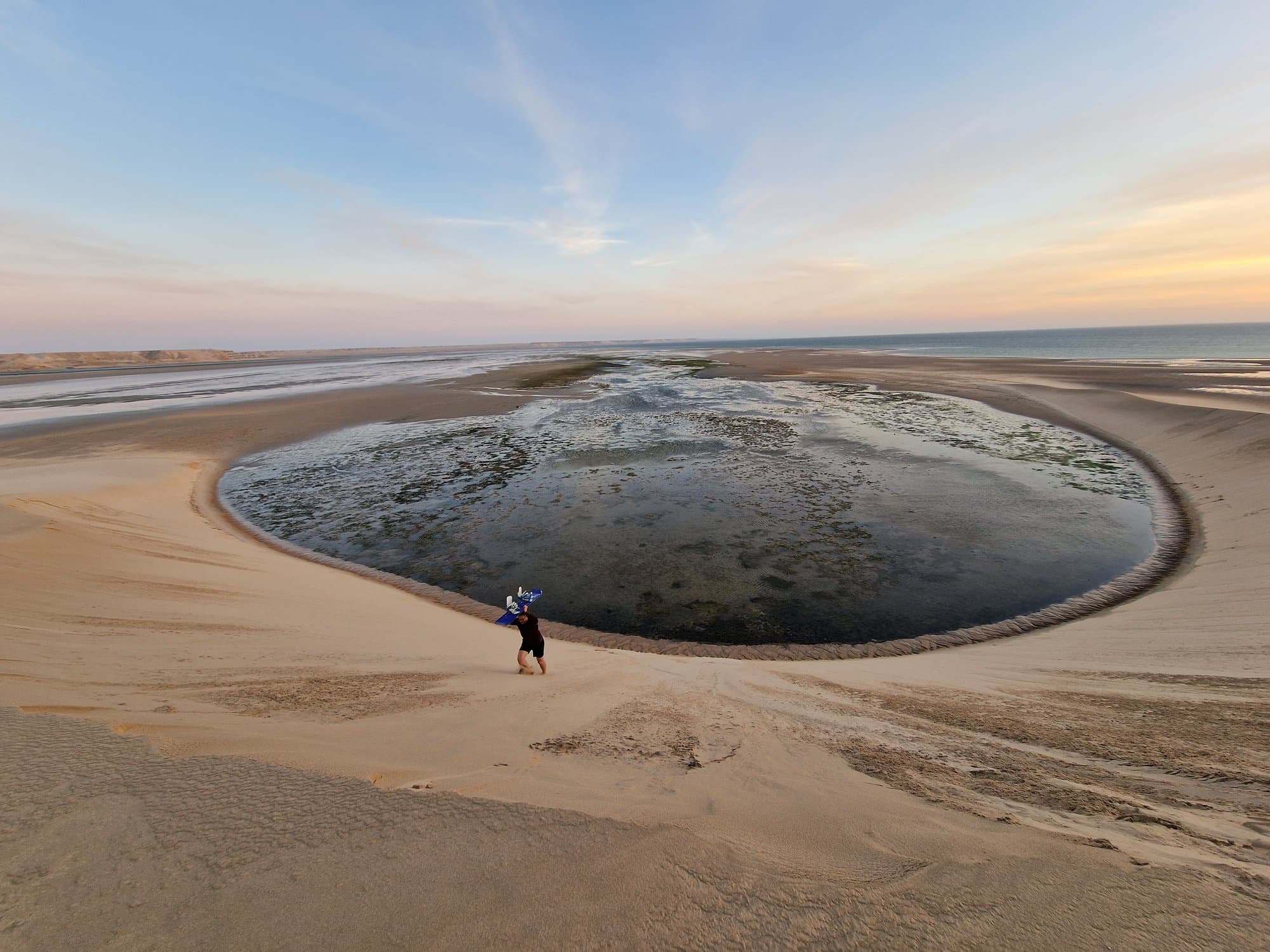 A horseshoe tidal pool ringed by white sand beneath the dune near Dakhla, a lone figure at its rim