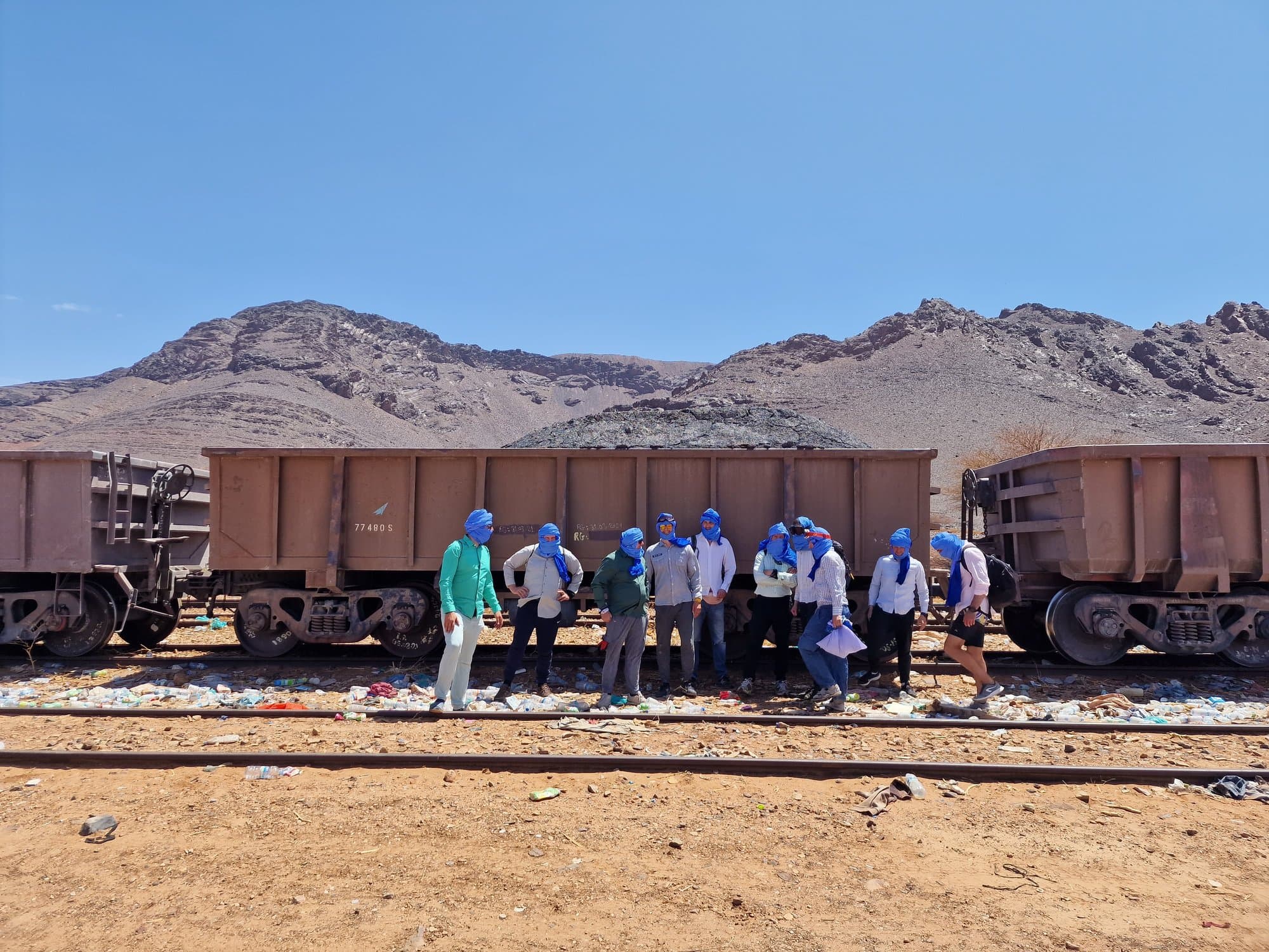 Travellers in blue headscarves lined up along empty iron-ore wagons at Zouerat