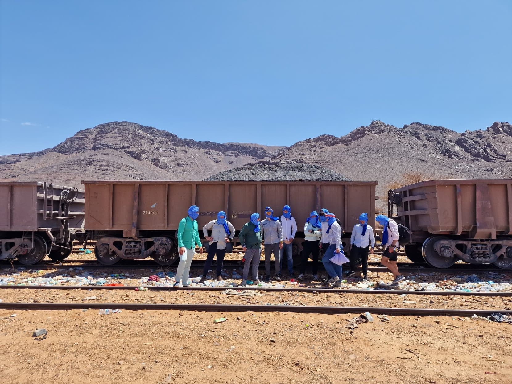 Travellers in blue headscarves lined up along empty iron-ore wagons at Zouerat